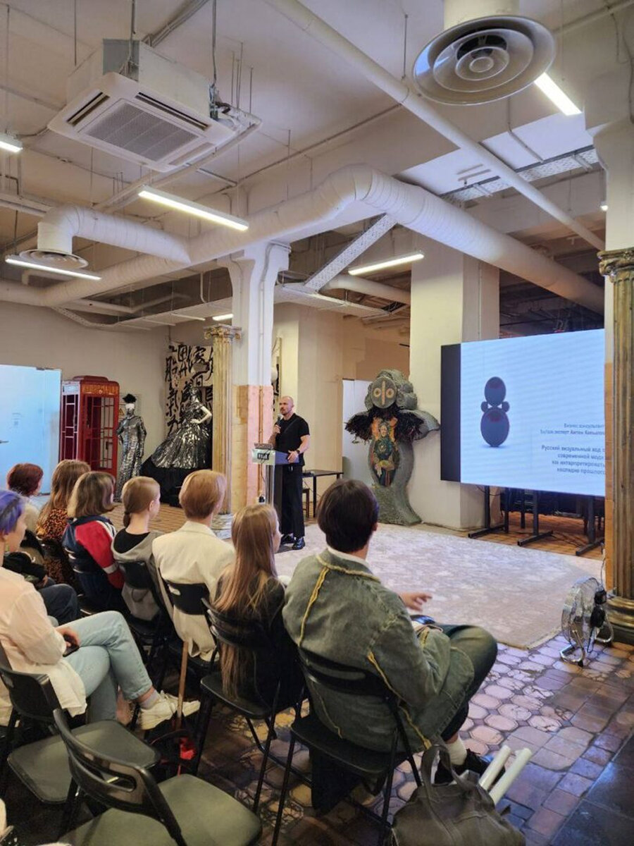 Anton Kopylov lecturing with a microphone to a seated audience in a loft space, slide on screen behind him