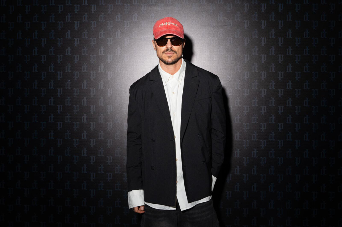 Anton Kopylov in a black double-breasted blazer, red cap and sunglasses against a press wall