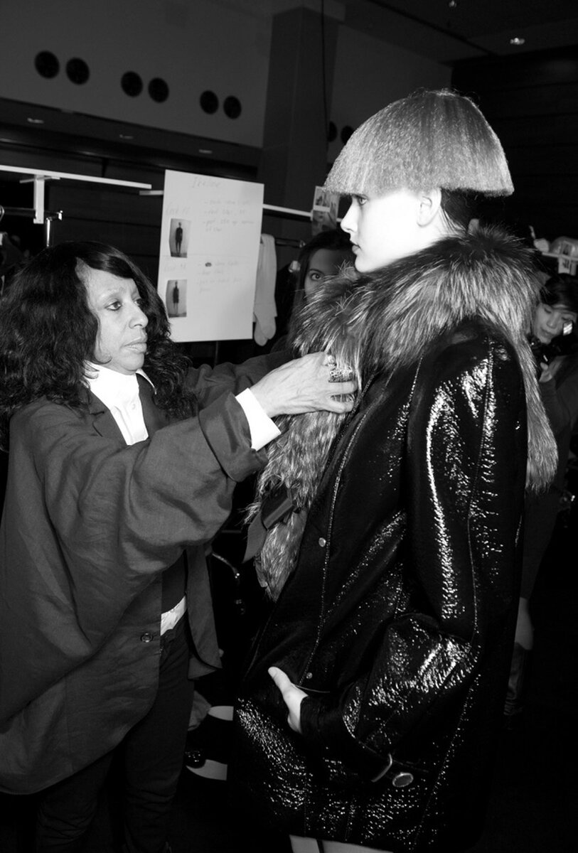 Backstage: a stylist adjusting a fur collar on a model in a patent leather coat and bowl-cut wig