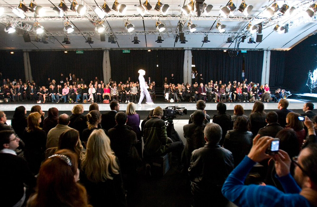 Wide view of a runway show — packed front row, lighting rig and a model in white mid-walk