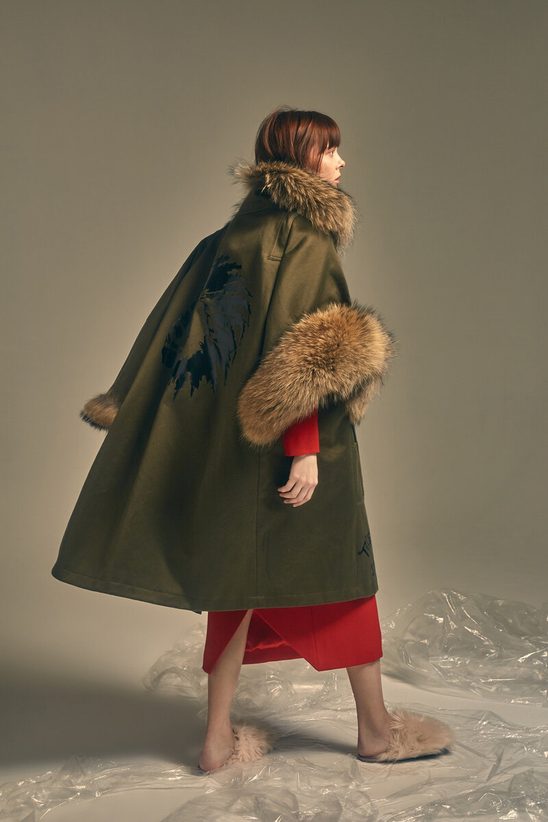 Model wearing khaki cape coat with fur collar and cuffs over a red dress, seen from the back