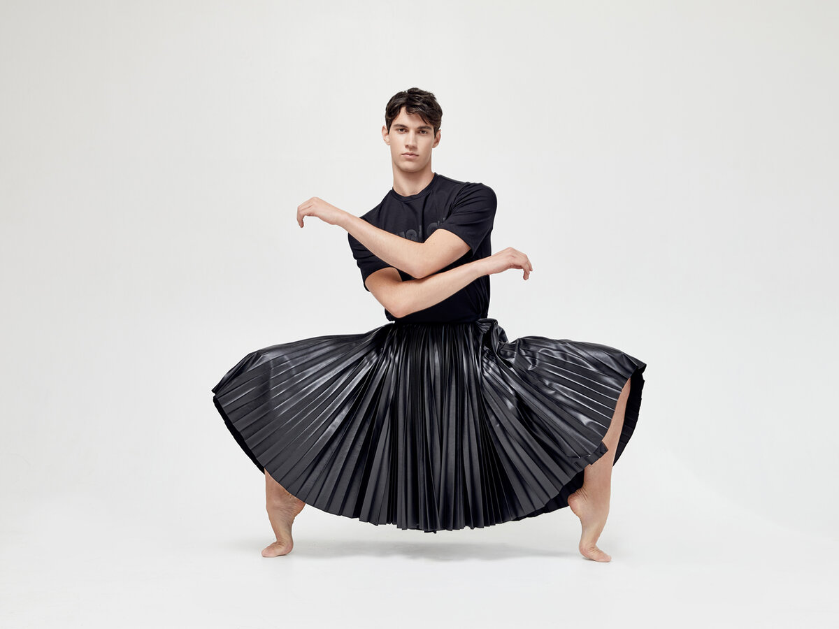 Male model in a black tee and flared pleated leather skirt, crouching barefoot