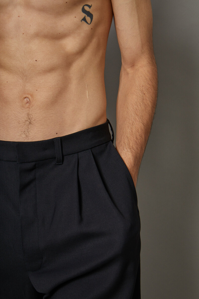 Close crop of black pleated wool trousers on a shirtless male model with S tattoo