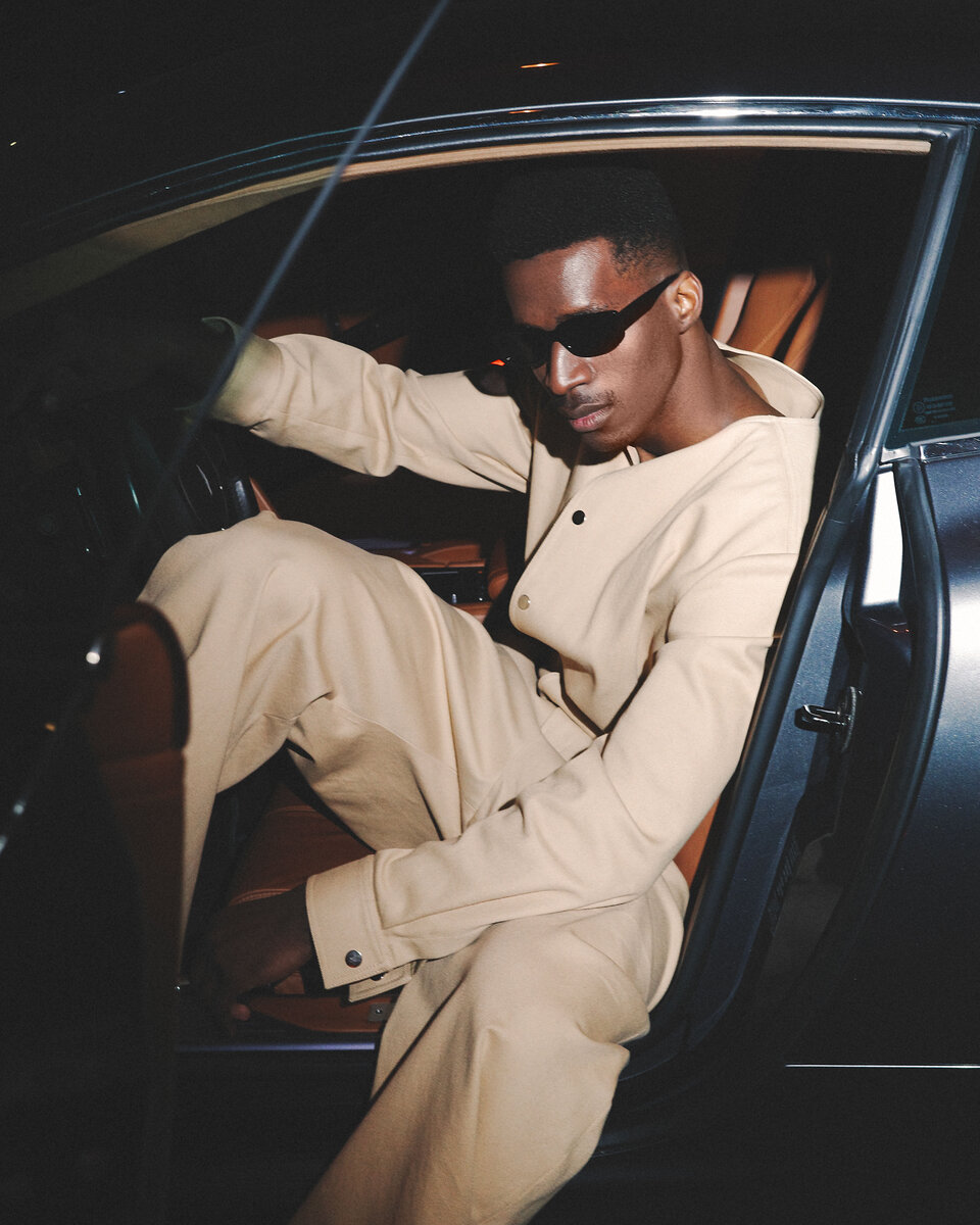 Black male model wearing beige outfit shot getting out of a front seat of a car